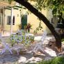 A table with a lemon tablecloth and two white chairs on a terrace under a tree.