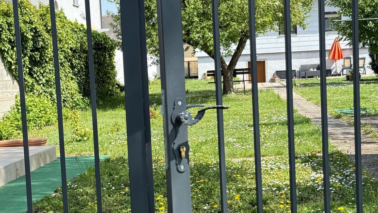 A garden with a metal gate in the foreground, behind it a tree and a house with a red parasol.