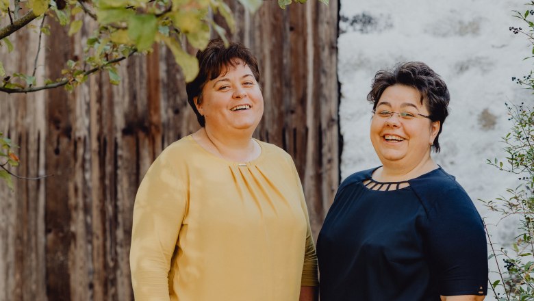 Two smiling women stand in front of a wooden wall outside.