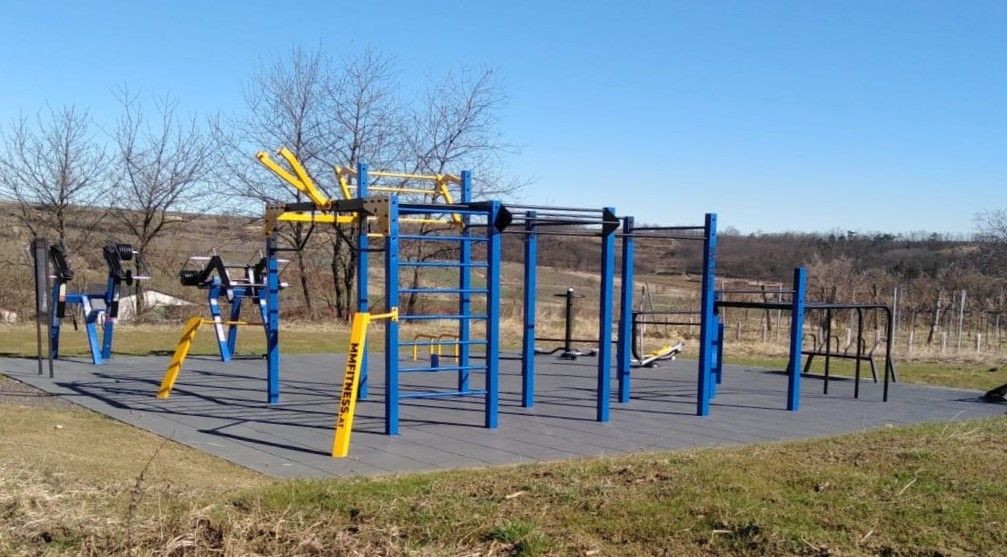 An outdoor calisthenics park with blue and yellow equipment, surrounded by a rural landscape.