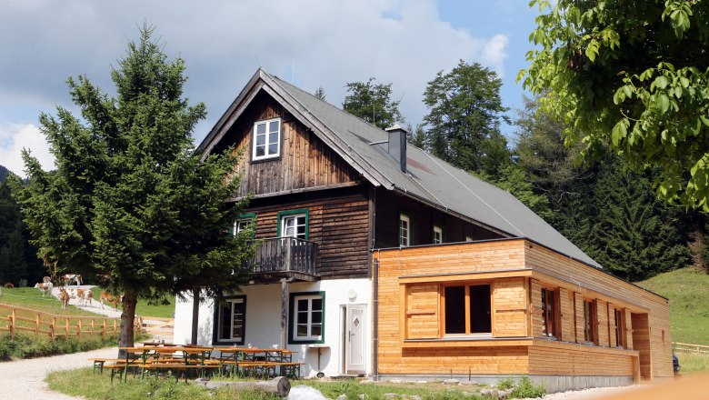 A traditional wooden house with extension in a rural setting, surrounded by trees and meadows.