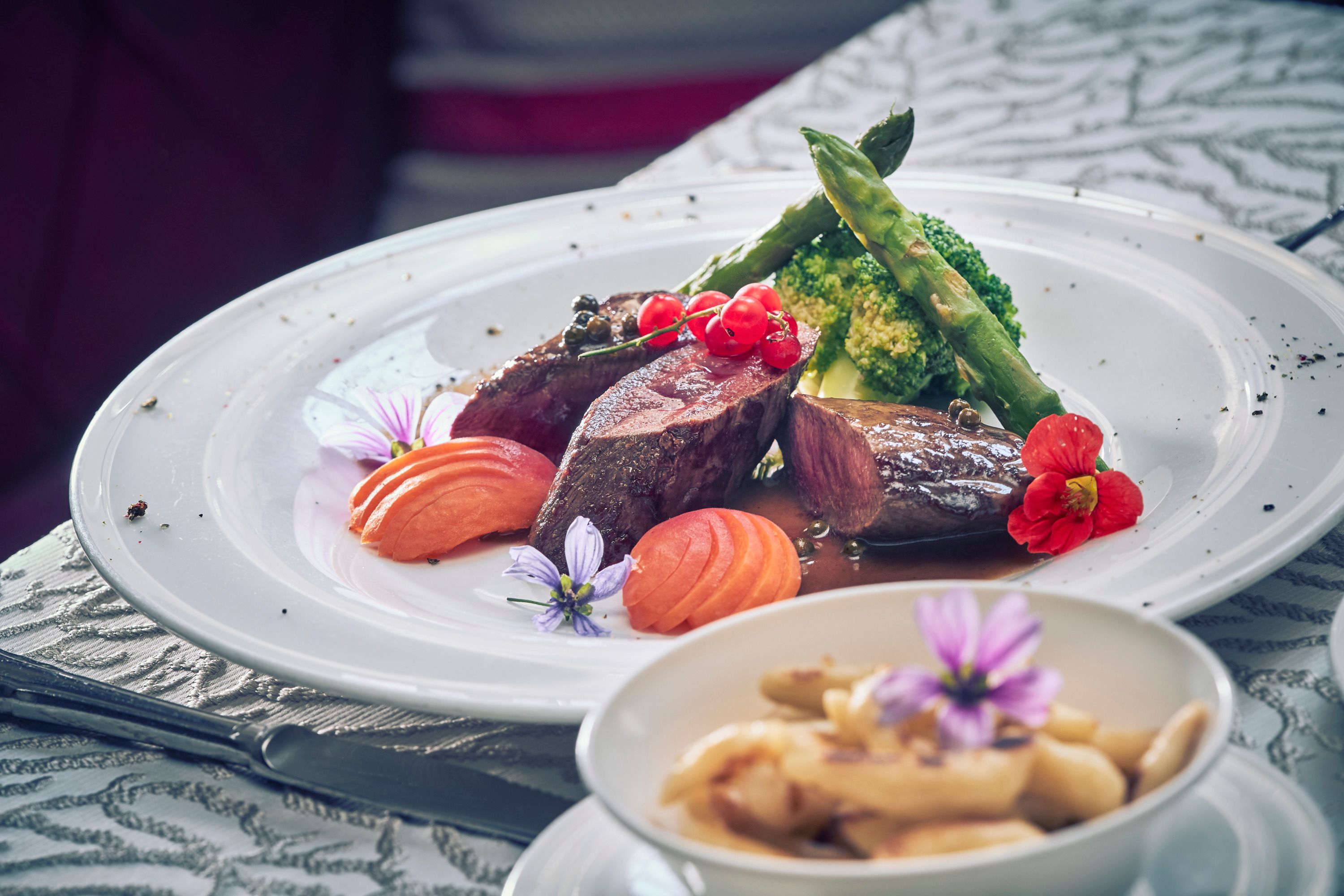 A plate of saddle of venison, peppery apricots, broccoli and asparagus, decorated with flowers. In the foreground, a bowl of potato noodles.