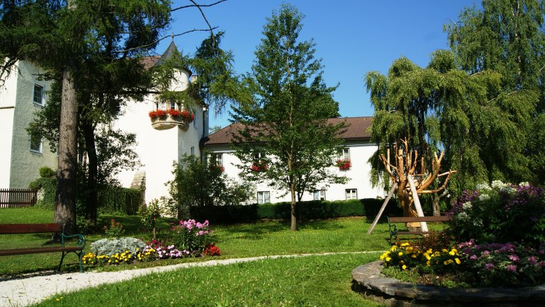 A castle with a white fa&ccedil;ade and red flowers, surrounded by a well-tended garden with trees and flowerbeds.