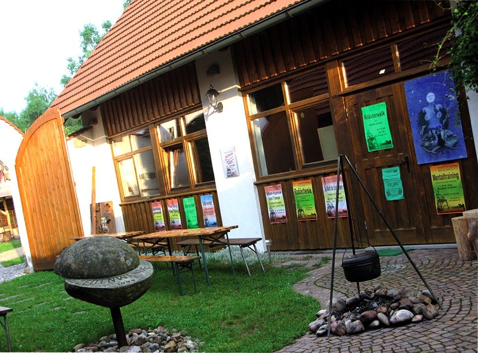 A courtyard with wooden tables, posters on a wooden wall and a cauldron over a fireplace.