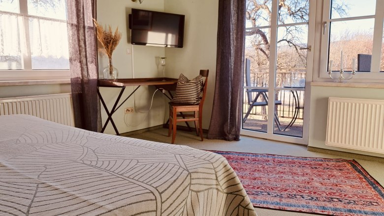 Comfortable room with bed, table, chair and TV. Large windows with a view of the terrace and trees. Carpet on the floor.