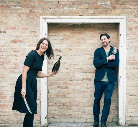 Two people in front of a brick wall, one holding a bottle and a knife, the other a glass.