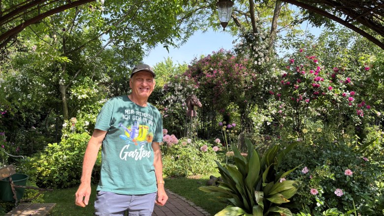 A man stands in a flowering garden under a pergola.