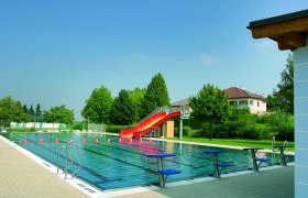 An outdoor pool with a swimming pool, a red slide and starting blocks. Trees and a house can be seen in the background.