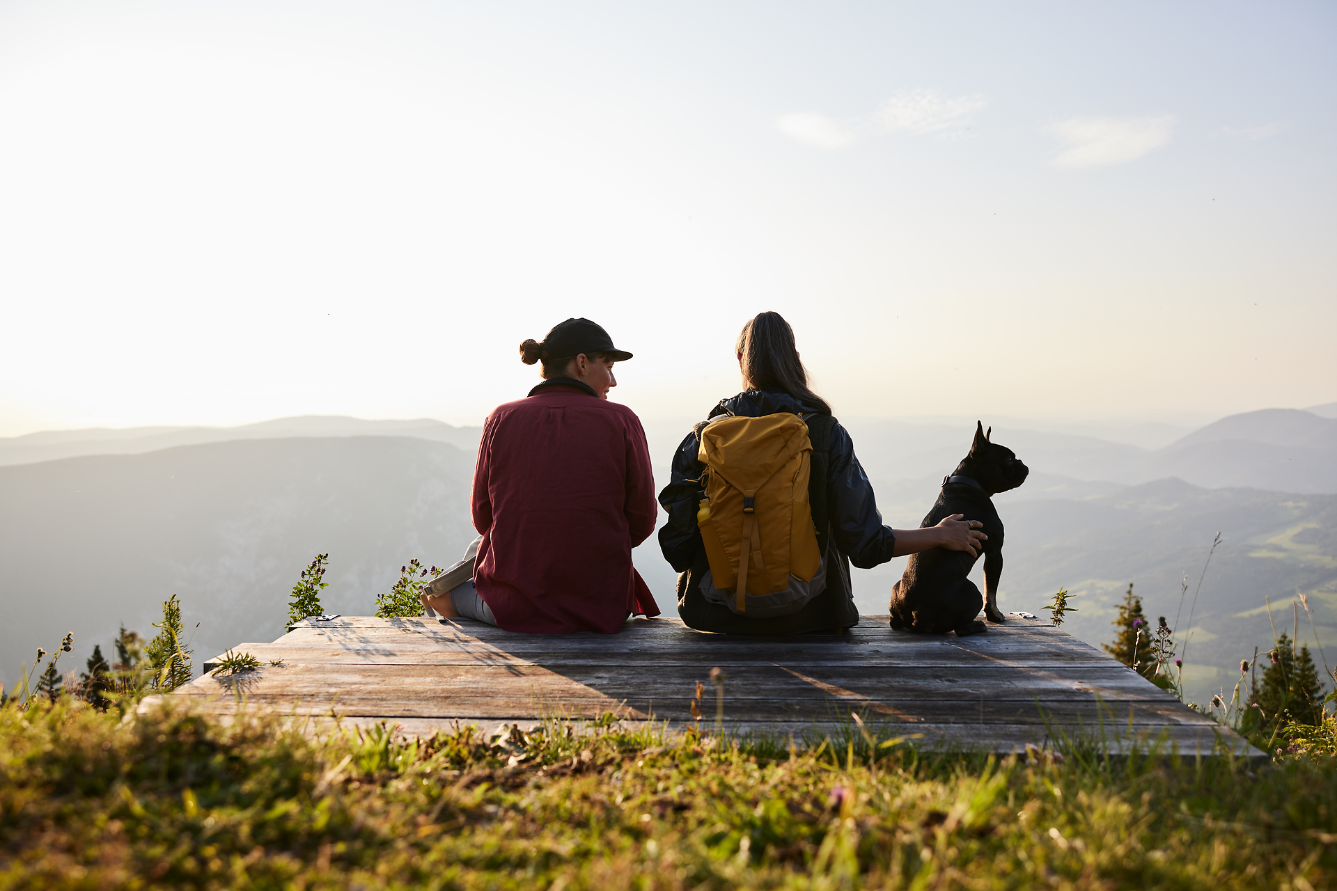 In the golden evening sun, two hikers enjoy the breathtaking view of the rolling hills and the wide valley. Their faithful companion, a small dog, sits relaxed next to them and shares this peaceful moment of closeness to nature. The fresh mountain air and the tranquillity of the surroundings invite you to unwind.