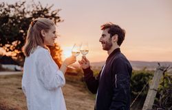 In der warmen Abenddämmerung stoßen zwei Menschen mit Gläsern an und genießen den Moment der Zweisamkeit. Umgeben von sanften Hügeln und Weinreben strahlt die Atmosphäre eine einladende Gemütlichkeit aus, die zum Verweilen einlädt.