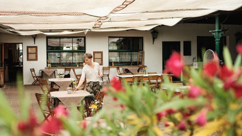 A woman prepares tables in a covered outdoor area of an inn. Blurred flowers can be seen in the foreground.
