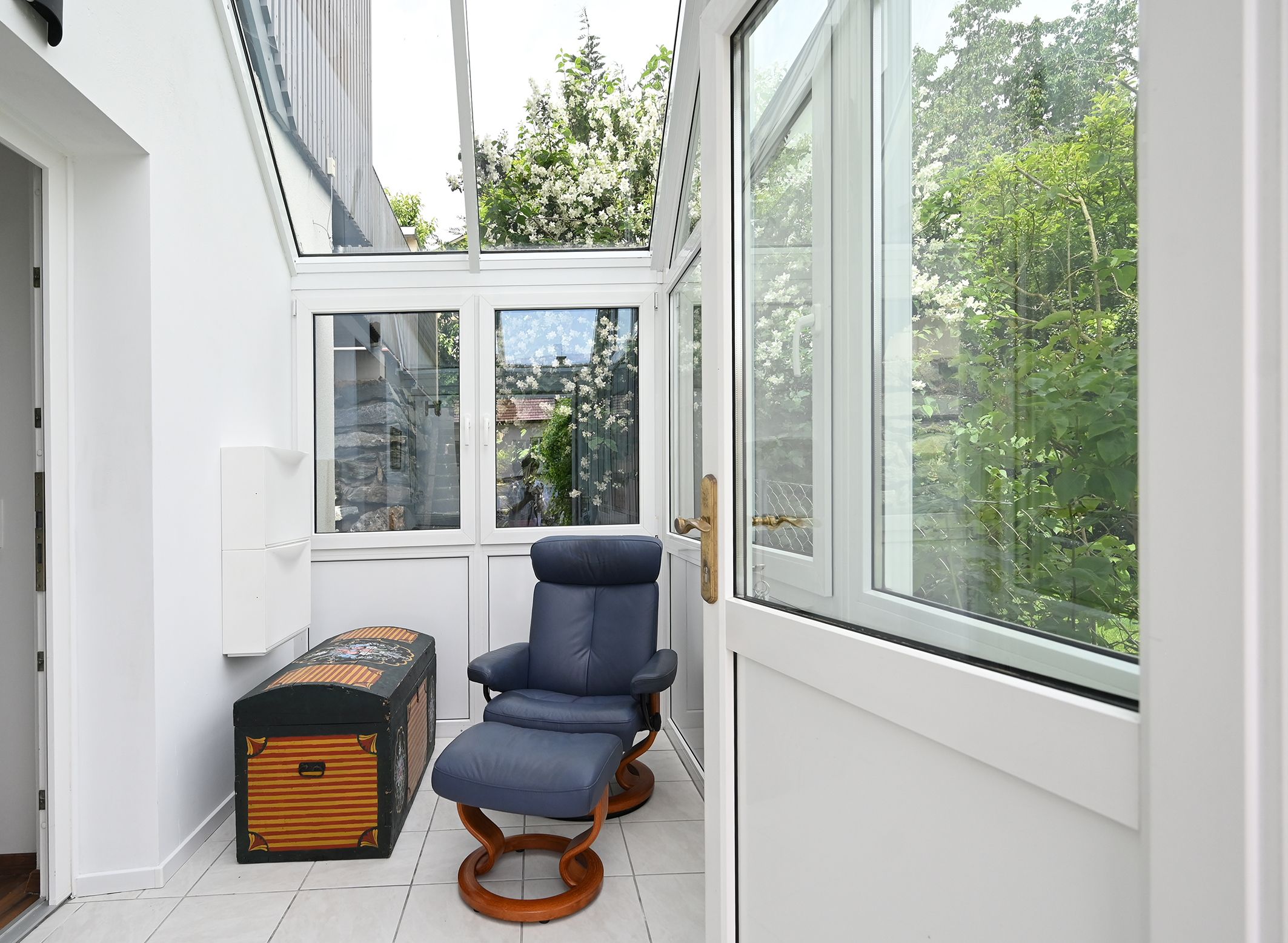 A bright anteroom with a blue armchair and a decorative chest, surrounded by large windows with a view of green plants.