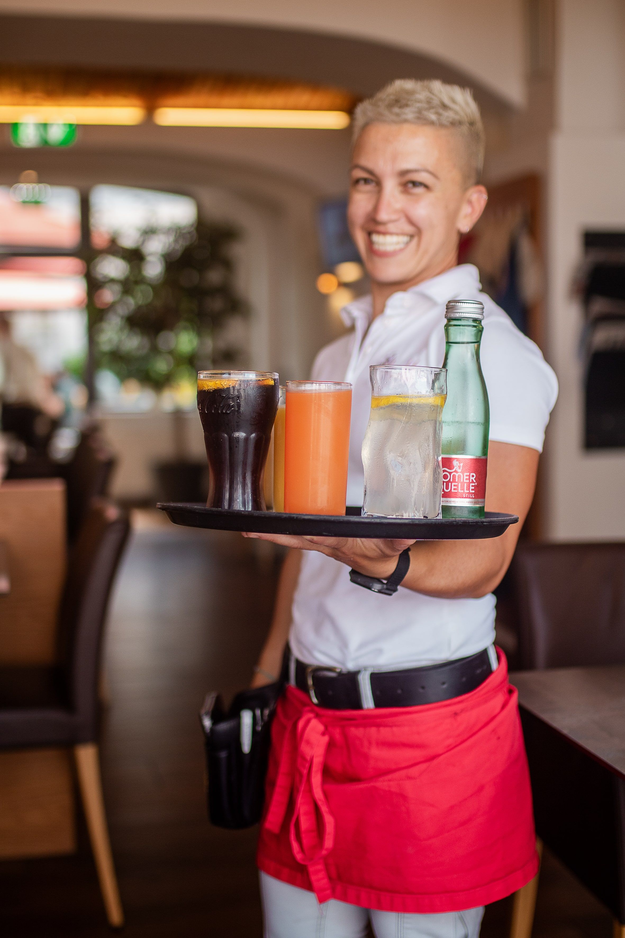 A smiling waitress serves drinks on a tray in a cozy restaurant.