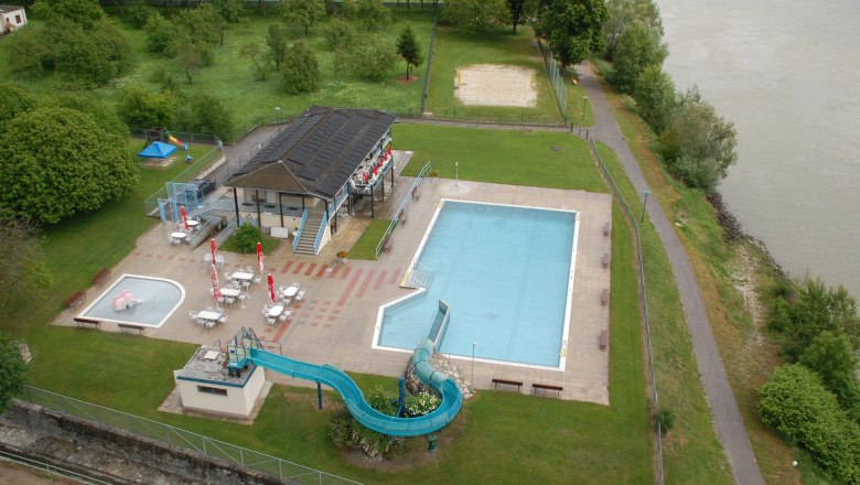 Aerial view of an outdoor pool with swimming pool, slide and sunbathing area on the riverbank.