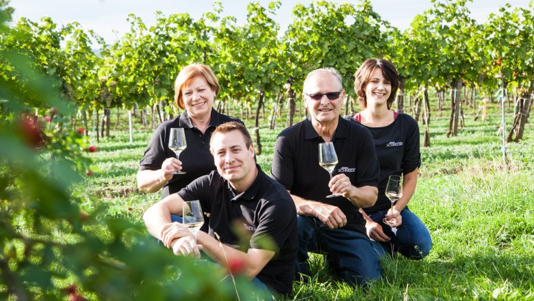 Four people are sitting in a vineyard holding glasses of wine. They are wearing black clothes and smiling at the camera.