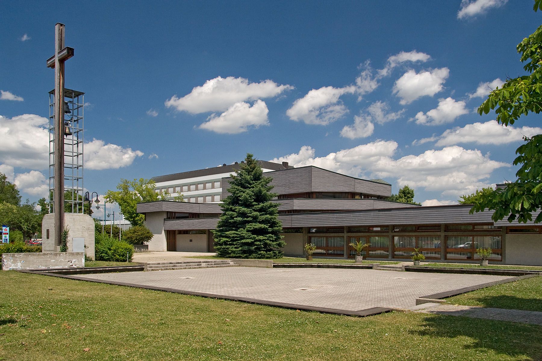 Modern church with bell tower and cross, surrounded by green spaces and trees, under a blue sky with clouds.