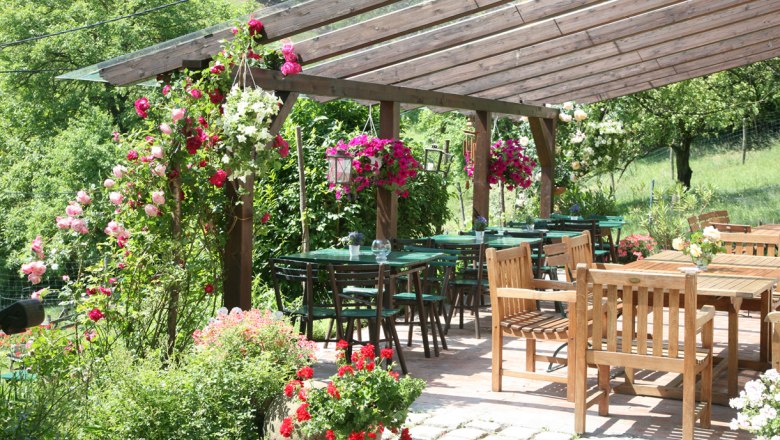 A cozy outdoor area of a wine tavern with wooden tables and chairs, surrounded by flowering plants and a wooden pergola roof.