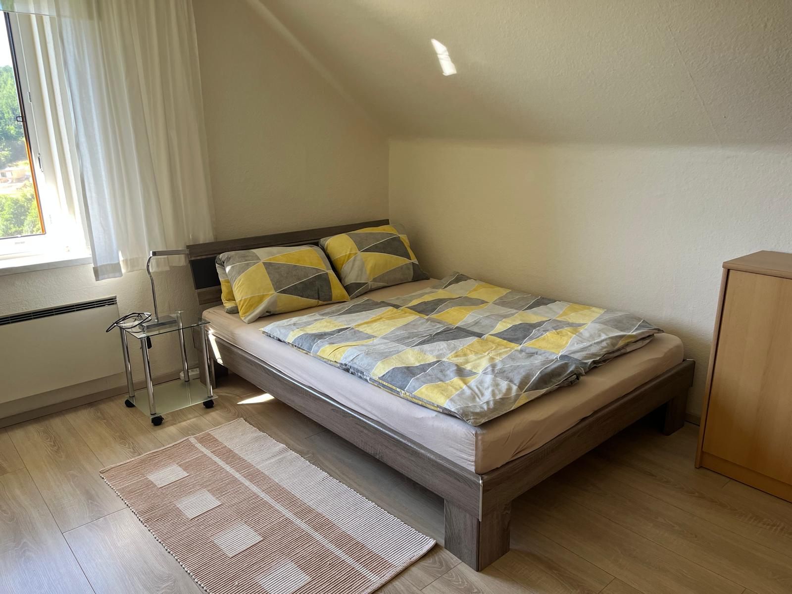 A cozy bedroom with a double bed, patterned comforter covers, a small bedside table and a window with curtains.