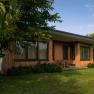 A modern wooden house with large windows, surrounded by trees and lawn.