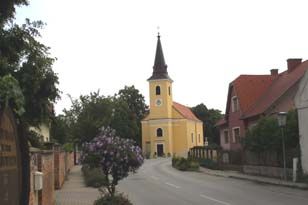 A yellow church with a tower in a quiet street, surrounded by trees and houses.