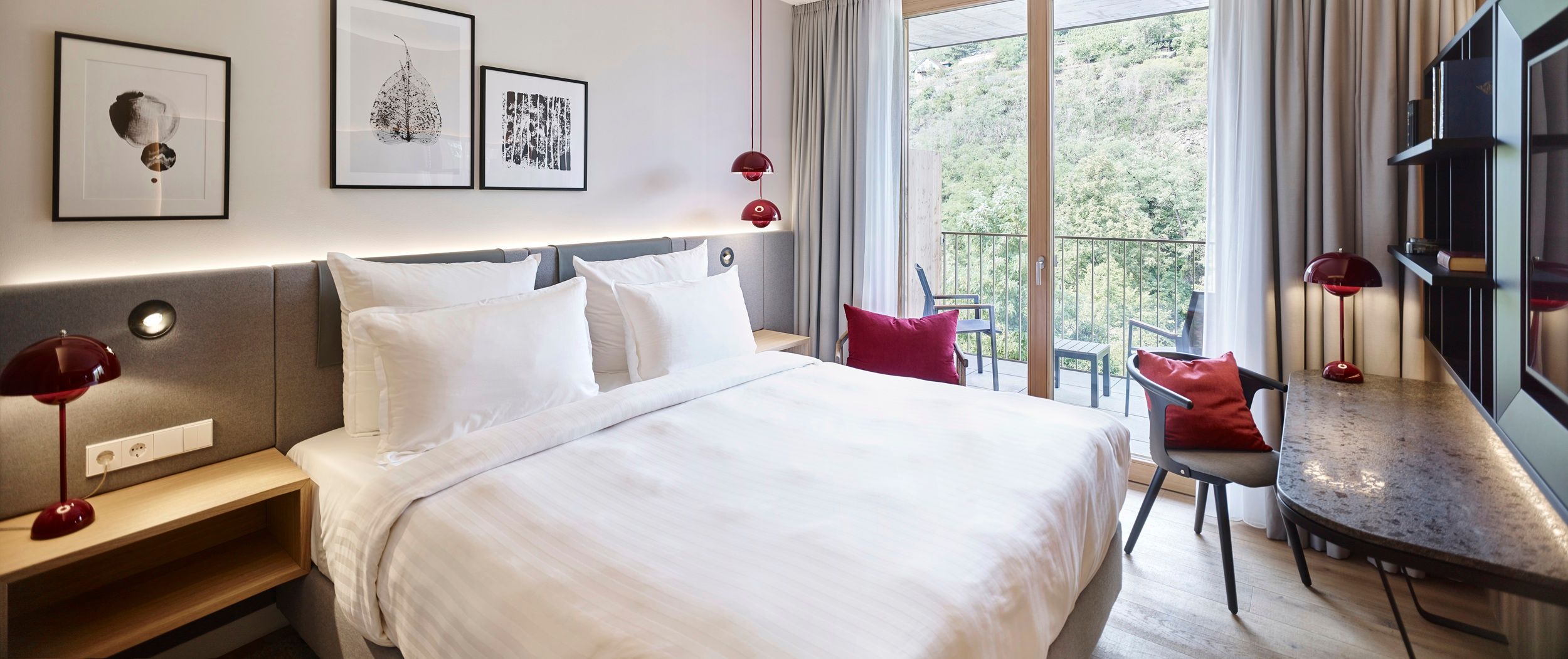Modern hotel room with large bed, desk and balcony with a view of the green landscape.
