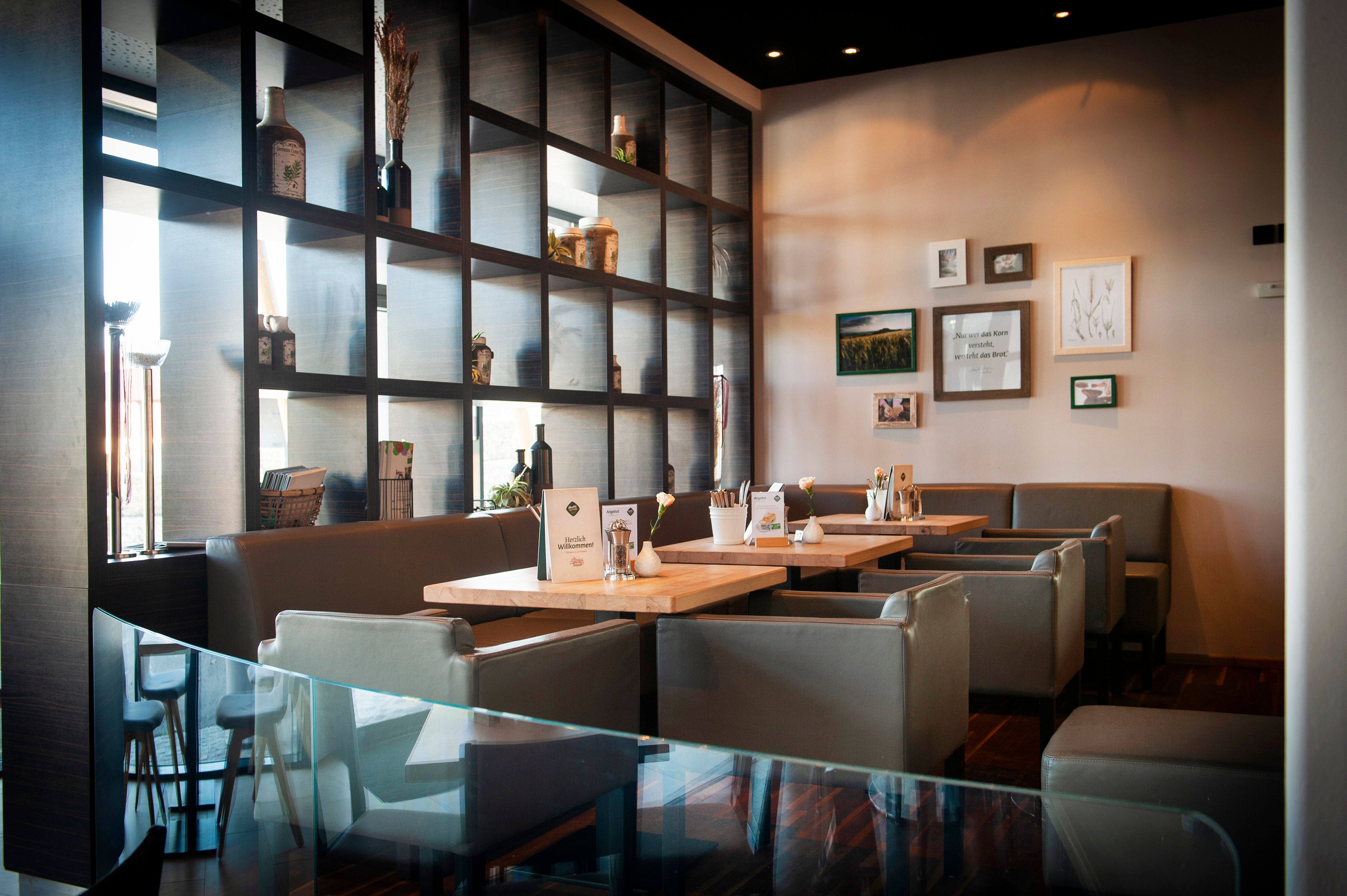 Interior view of a modern café with wooden tables and gray chairs, decorated with plants and pictures on the wall.