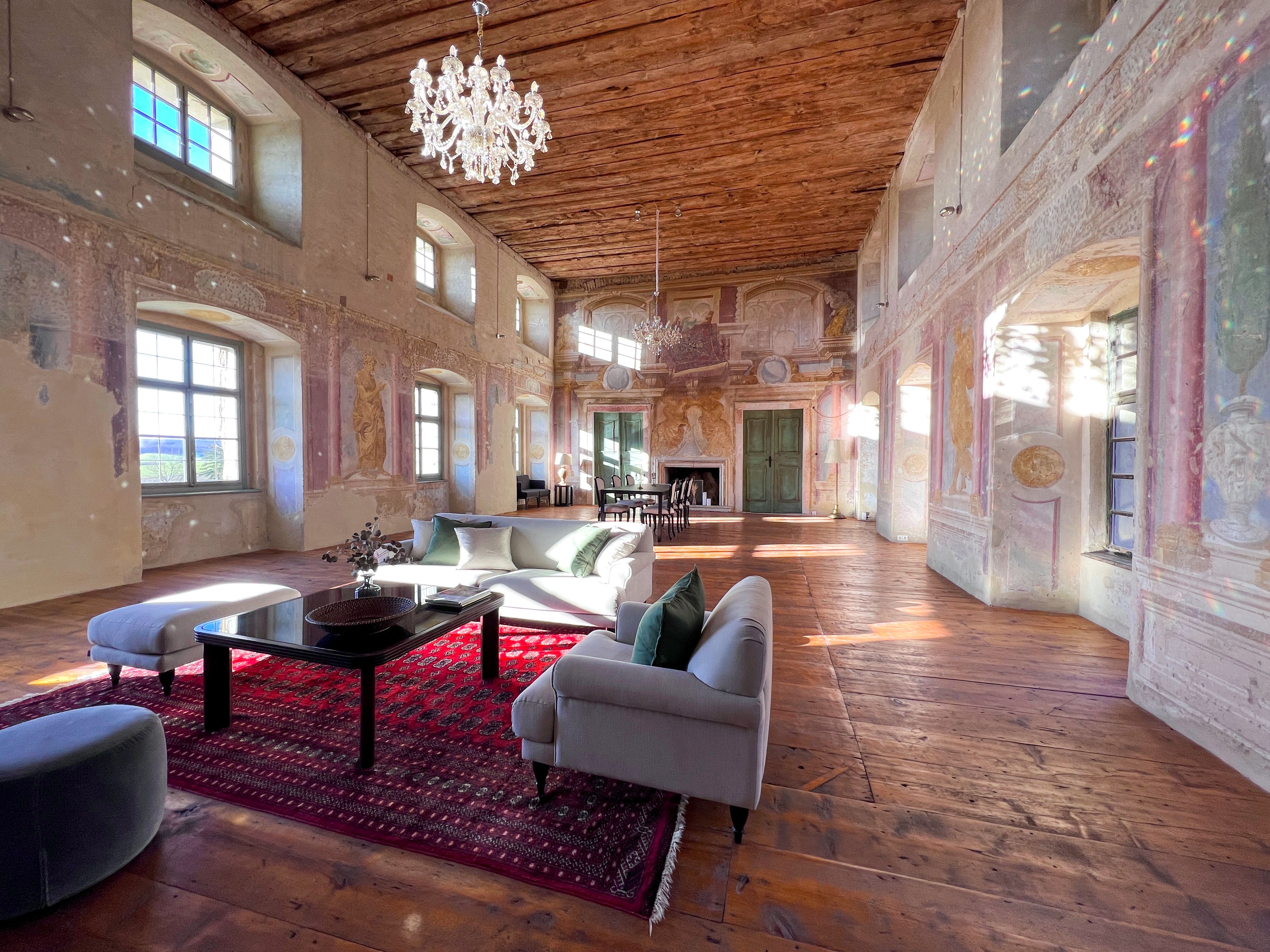 A historic ballroom with a wooden floor, painted walls and a chandelier. Modern furniture and a red carpet are placed in the room.