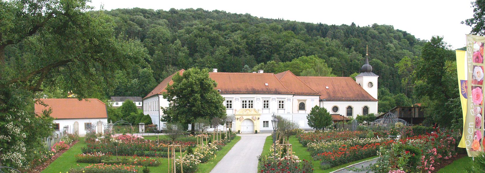 A historic building with a red roof, surrounded by a rose garden and trees, with a wooded hill in the background.