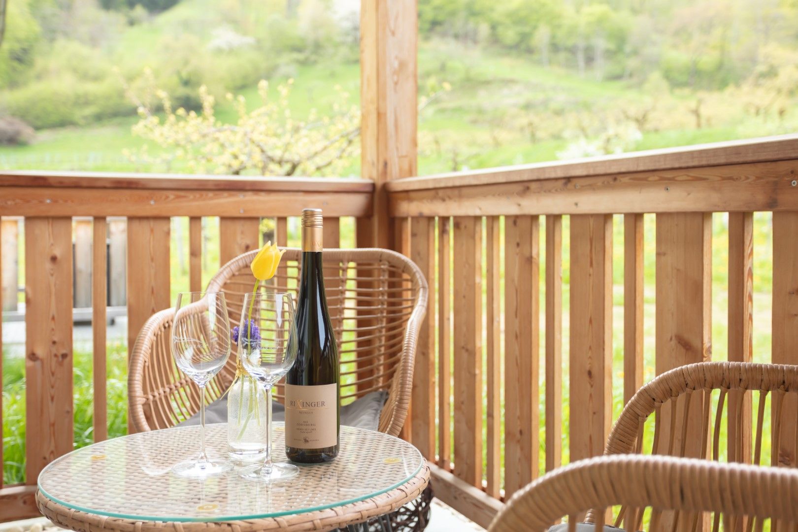 A balcony with wooden furniture, a bottle of wine and glasses on a table. Green landscape in the background.