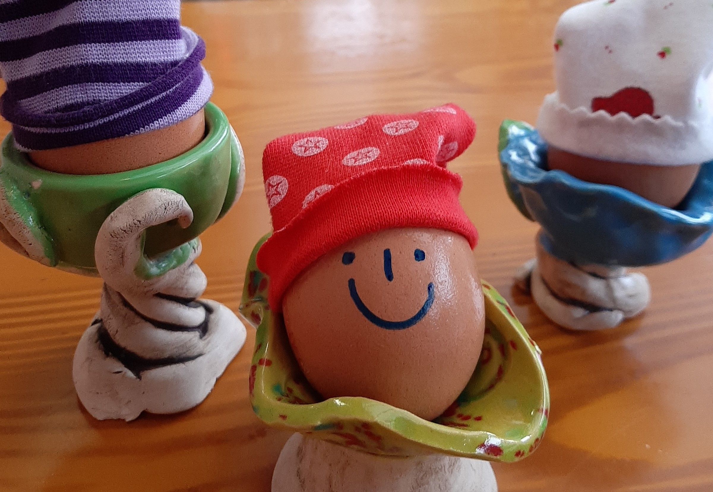 Three eggs in colorful egg cups, one with a painted face and a red cap.