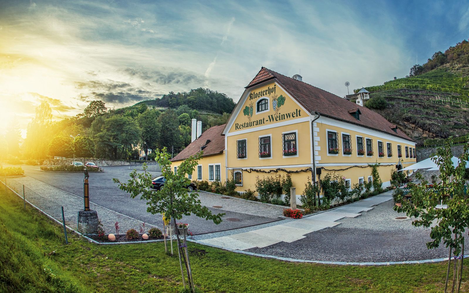 Exterior view of a yellow building with the inscription 'Klosterhof Restaurant-Weinwelt', surrounded by trees and vineyards at sunset.