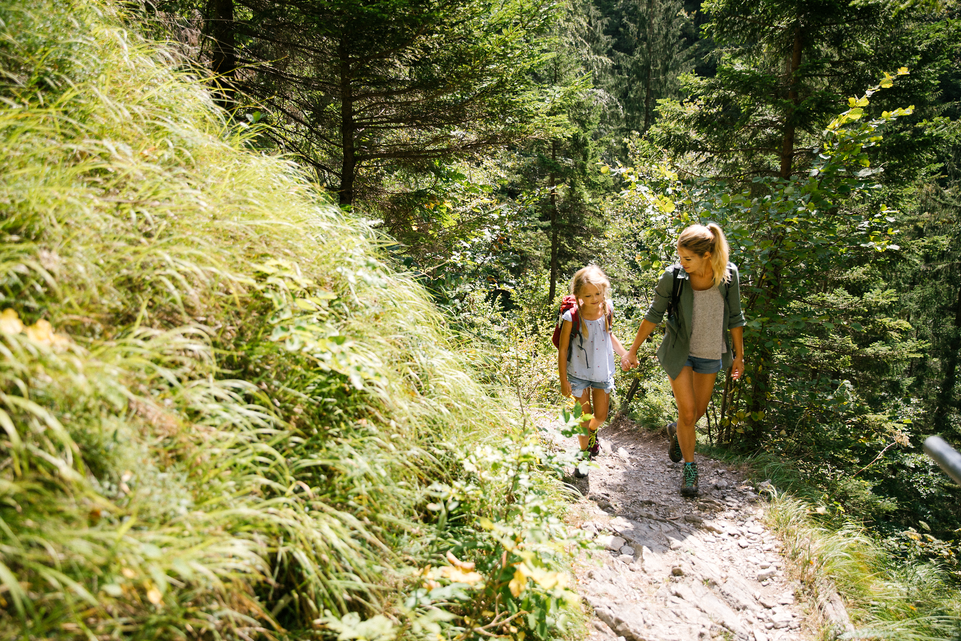 A mother and daughter walk happily along a narrow path surrounded by lush greenery and tall trees. The fresh air and the gentle babbling of a nearby stream create an inviting atmosphere for families who want to explore nature.