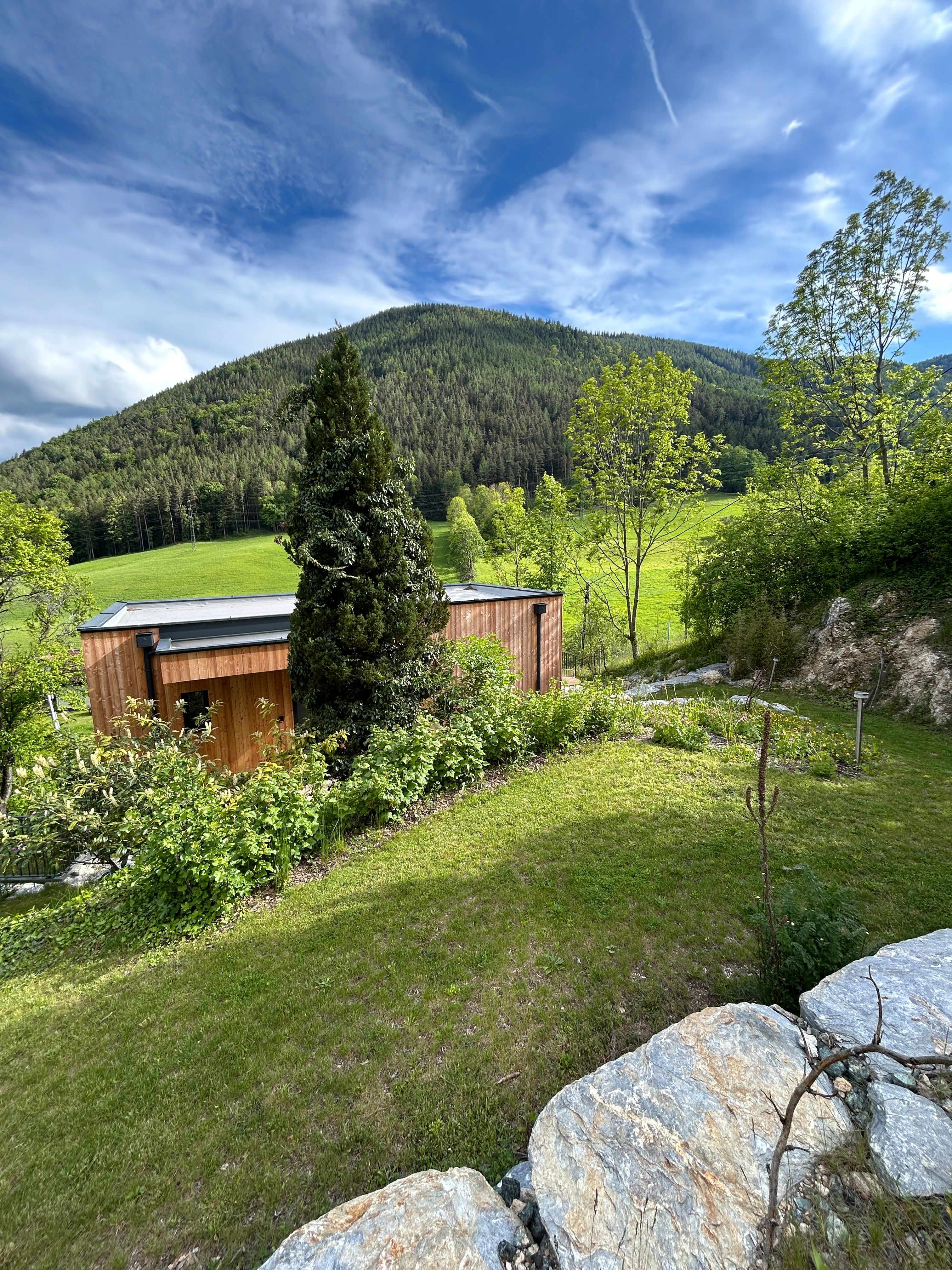 A small wooden house stands in a green landscape with a wooded hill in the background and a blue sky above.