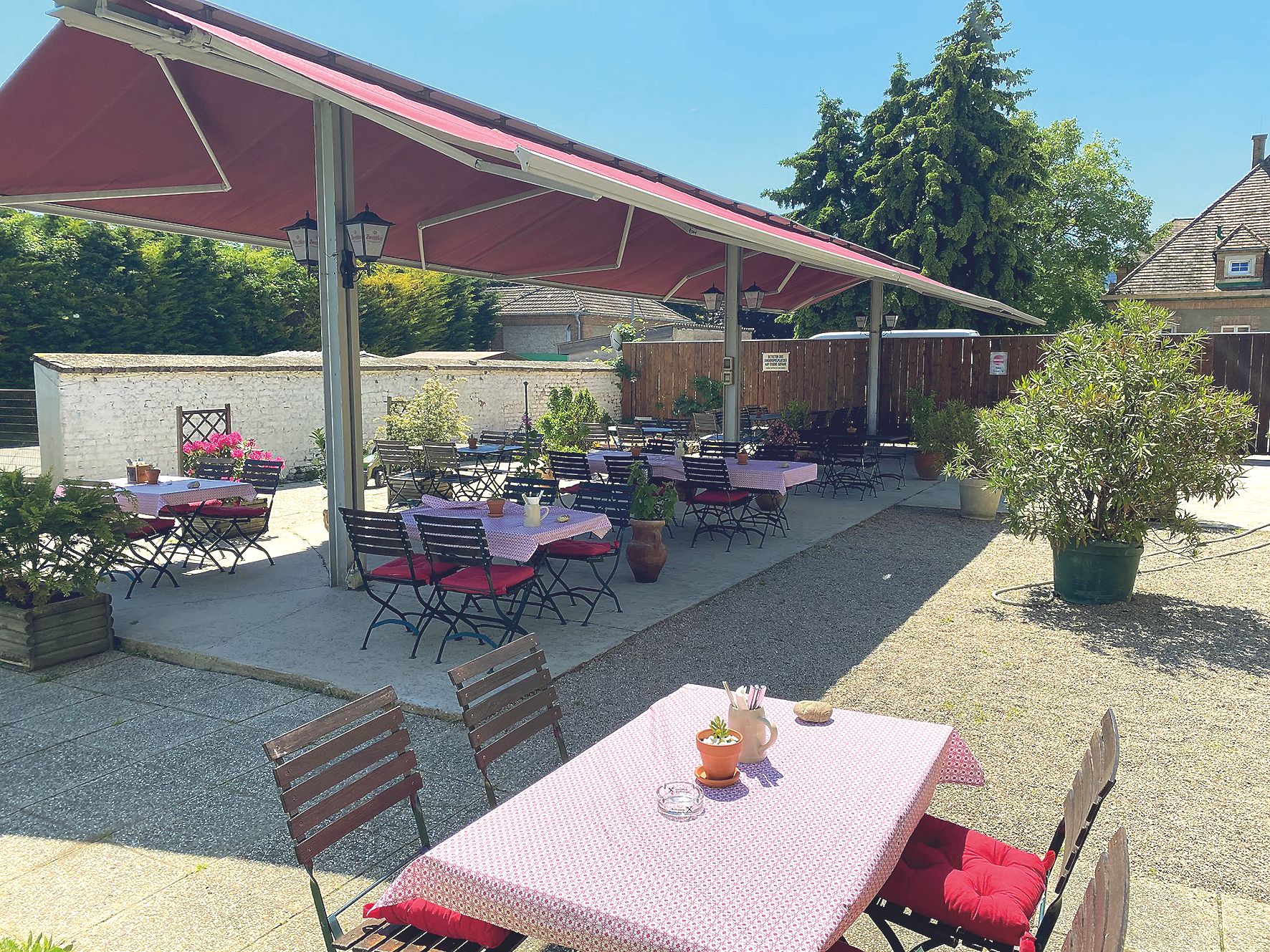 A guest garden with tables and chairs under a sunshade, surrounded by plants.