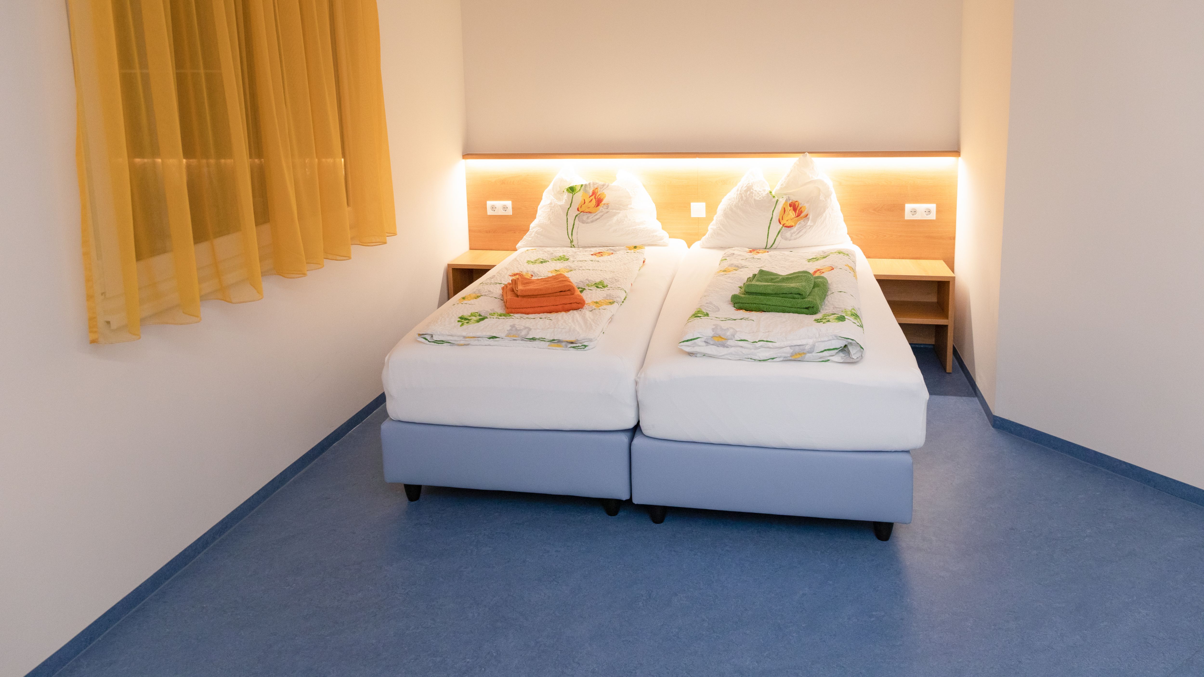 A double room with two single beds, yellow curtains and blue floor. The beds are decorated with floral bedding and colorful towels.