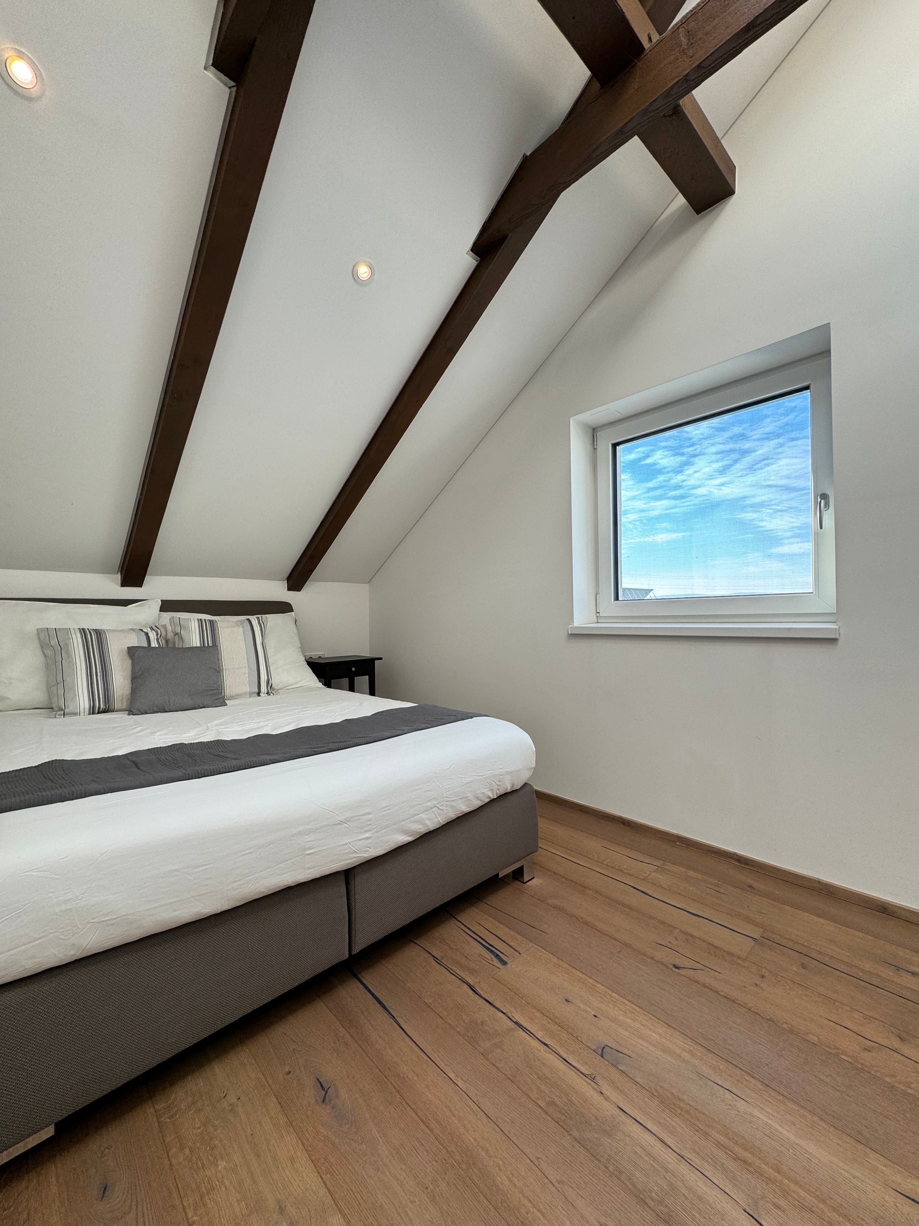 Modern bedroom with wooden beamed ceiling, large bed and window with a view of the blue sky.