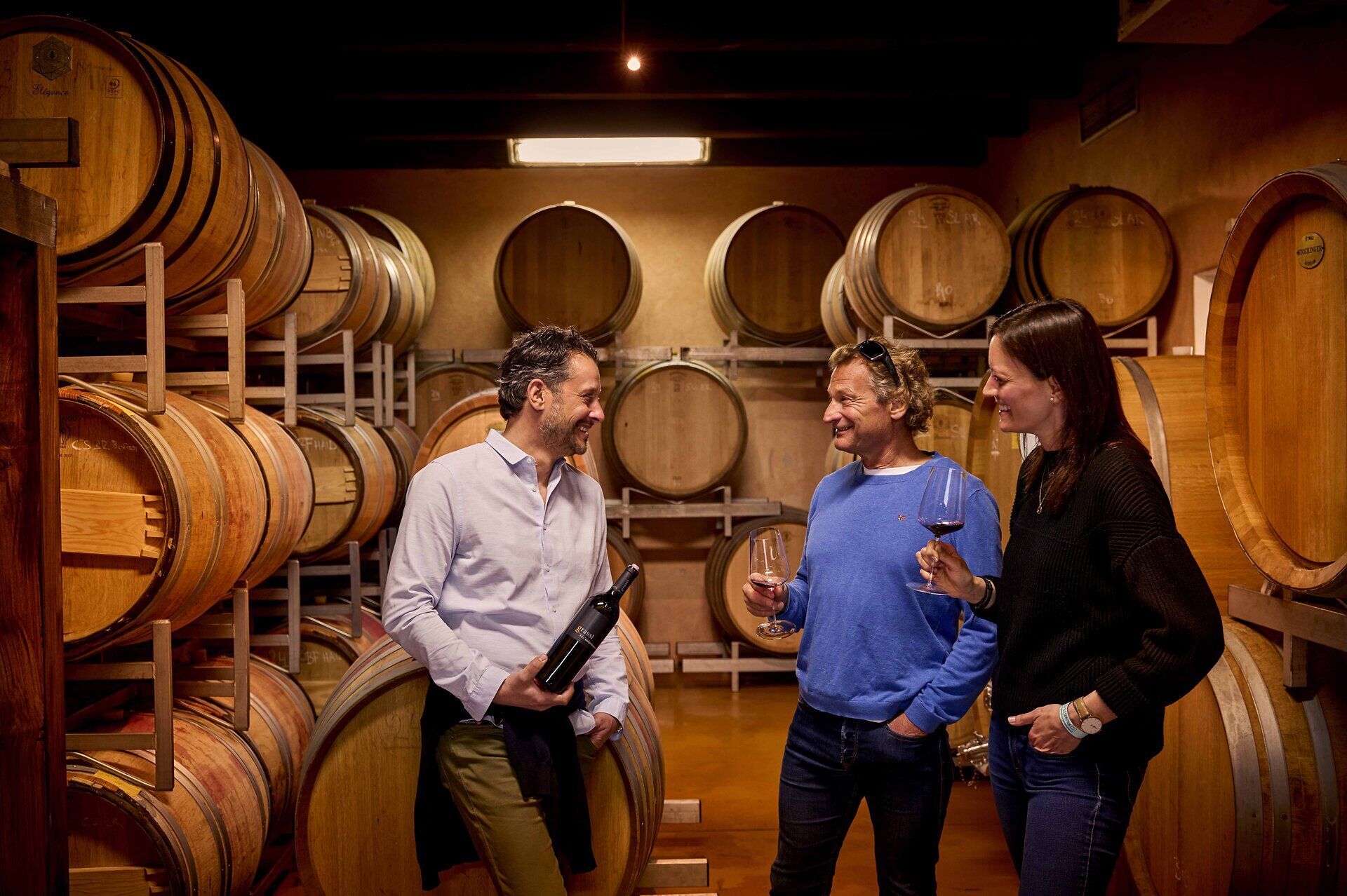 Philipp Grassl and two other people are tasting wine in his wine cellar. In the background, you can see several wooden barrels stacked on top of one another.