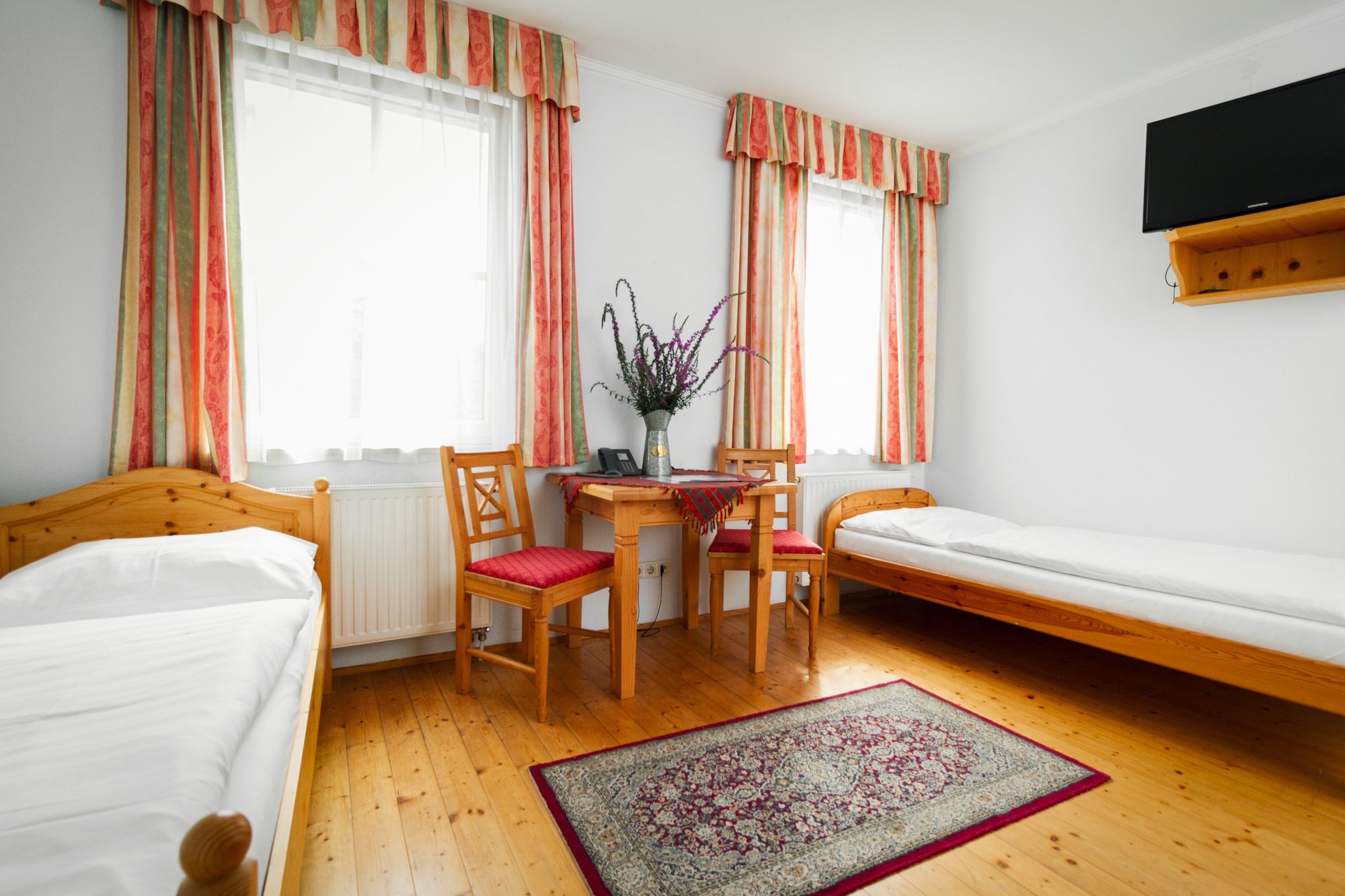 A simply furnished room with two single beds, a table with two chairs, a carpet and a TV on the wall.