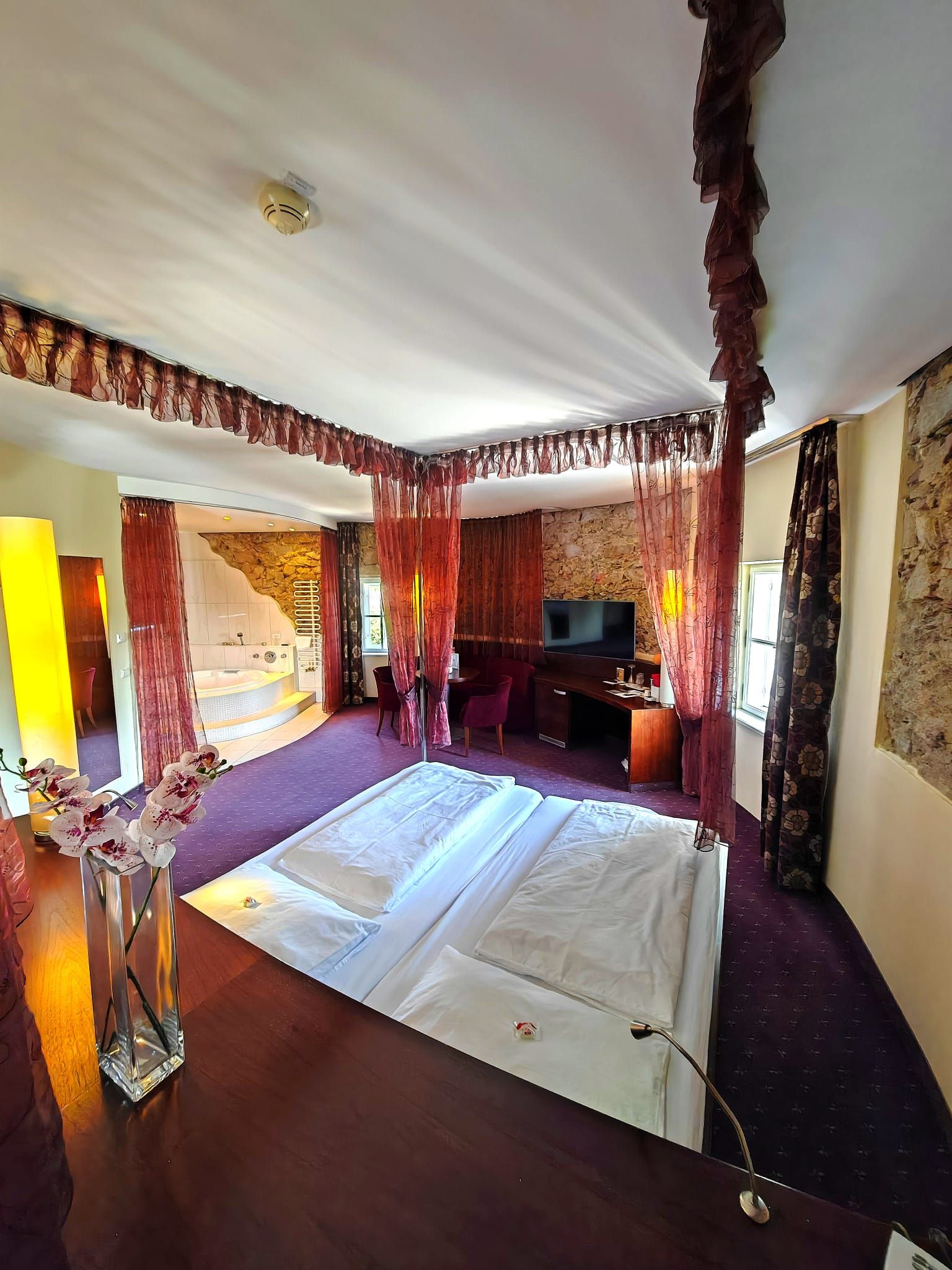 A luxurious hotel room with a large bed, red curtains, a television, and a bathtub in the background.
