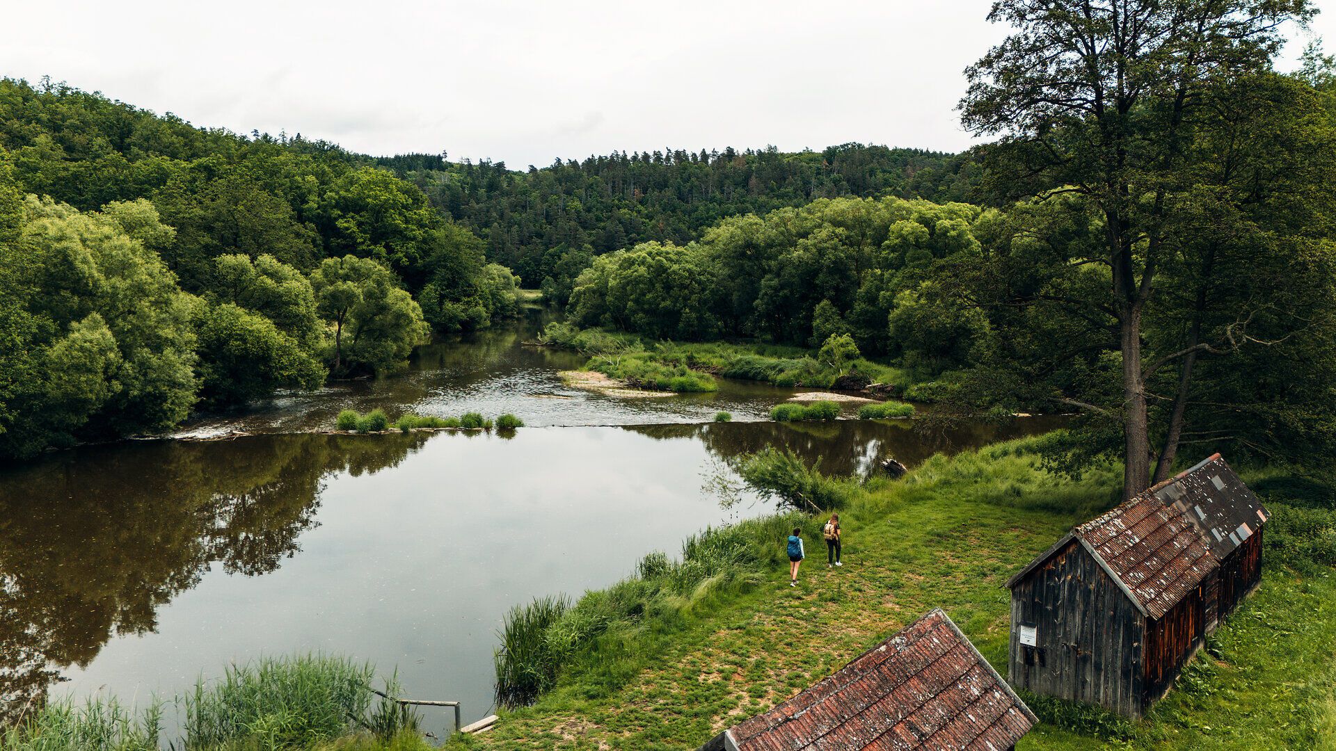 Two people walk along a riverbank, flanked by dense trees and old wooden huts, in Drosendorf in the Waldviertel.