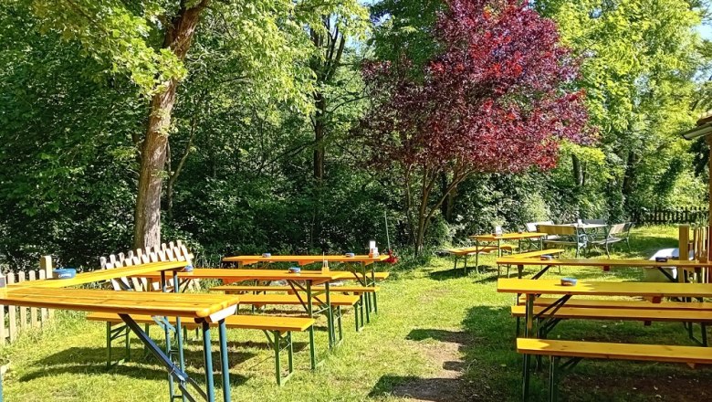 A beer garden with empty wooden benches and tables under trees.