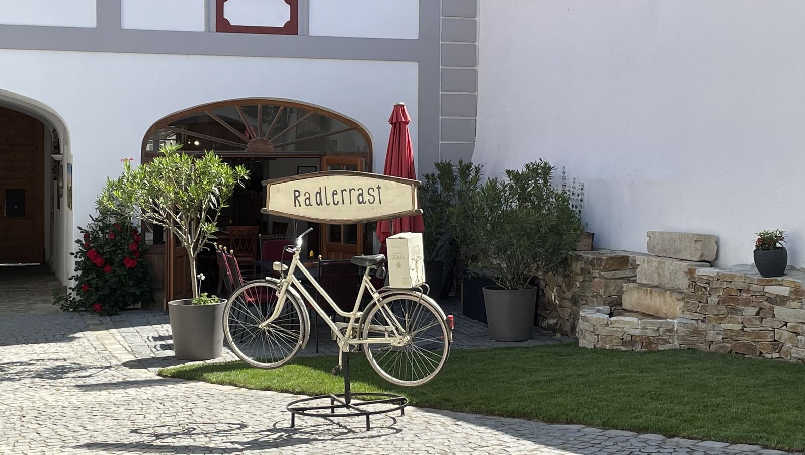 A decorative bicycle with a "cyclists' rest" sign stands in front of a building with plants and a parasol.