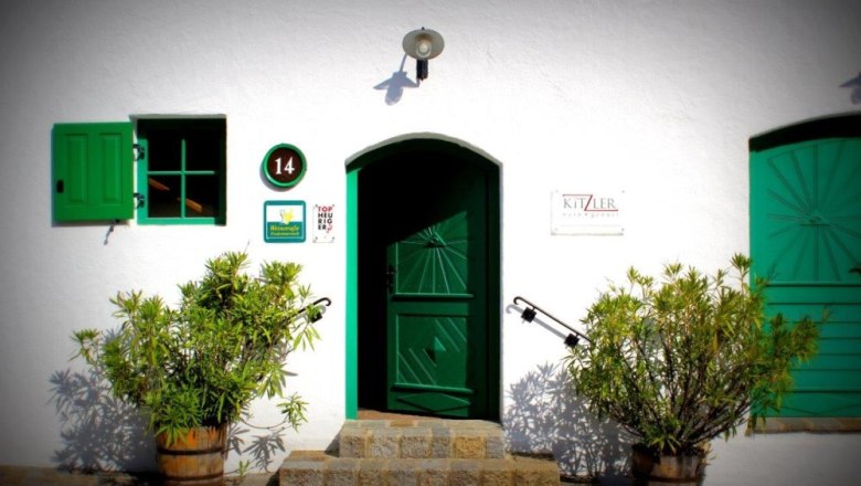 Entrance of a wine tavern with green door and shutters, surrounded by plants in wooden barrels.