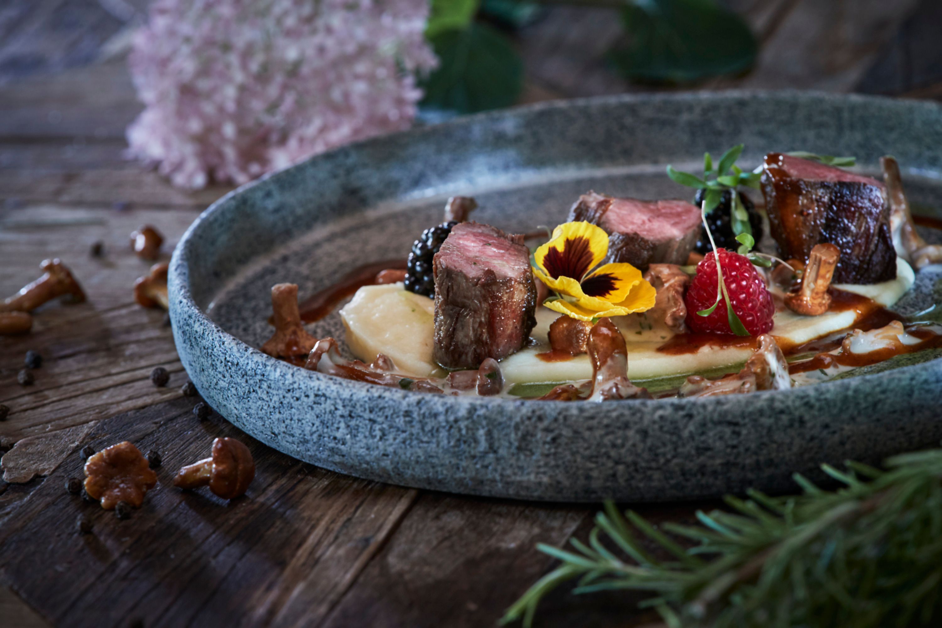 A plate of venison fillet, chanterelles, berries and edible flowers on a wooden table.