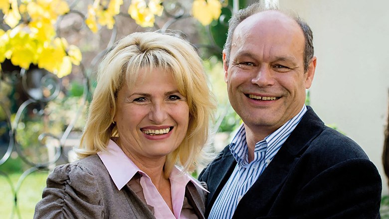 A smiling couple stands outside in front of a background of yellow leaves.