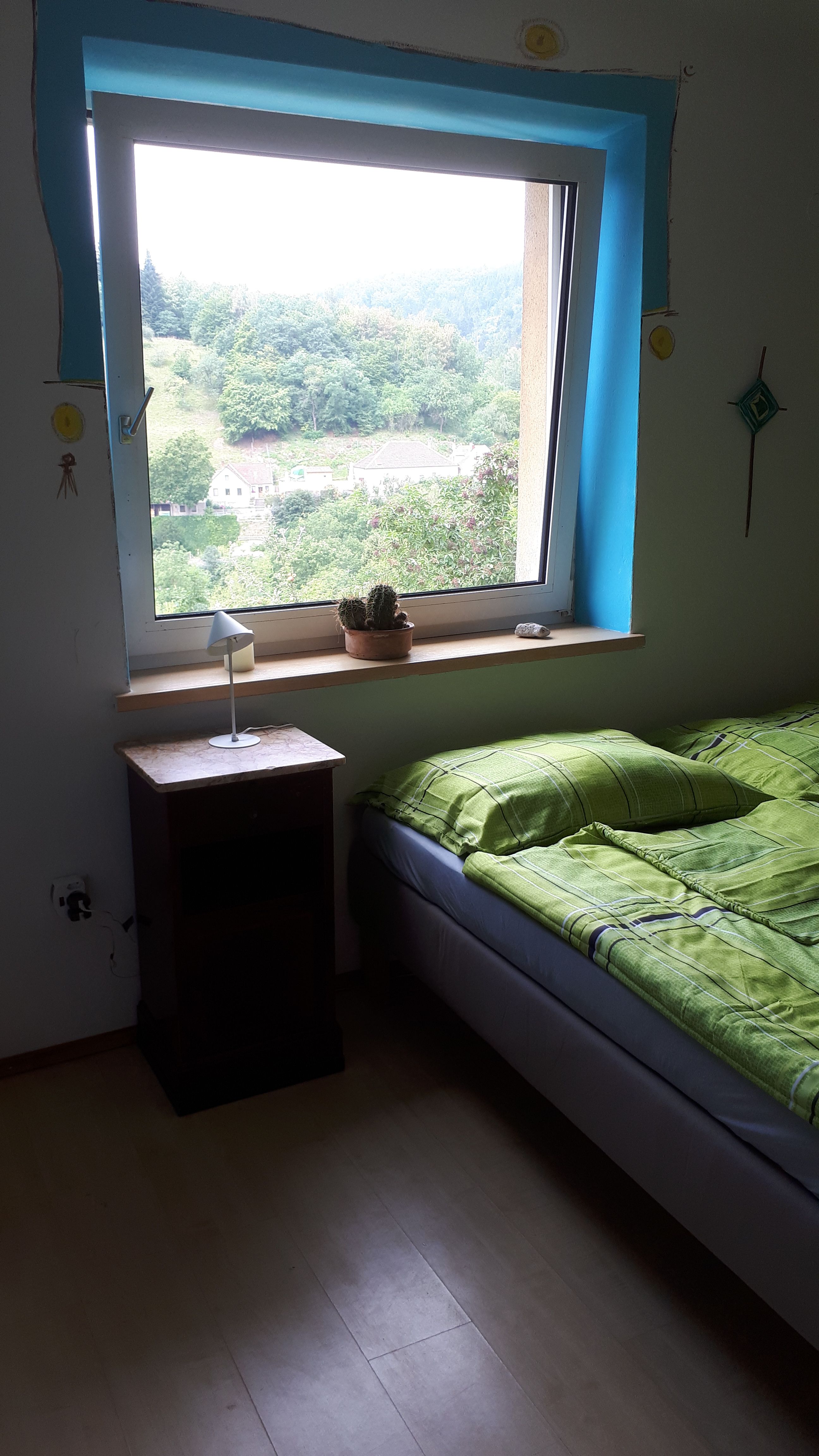A bedroom with a bed, a bedside table and a window overlooking a green landscape.