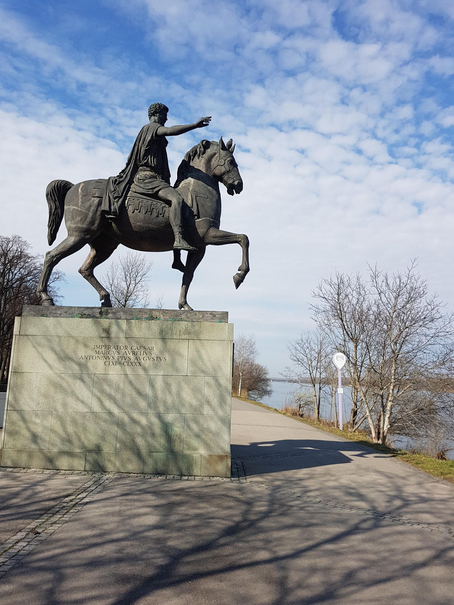 Equestrian statue of Marcus Aurelius on a pedestal, surrounded by trees and a path by the water.