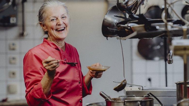 A smiling woman in a red chef's jacket stands in a kitchen with a spoon and a bowl in her hand.