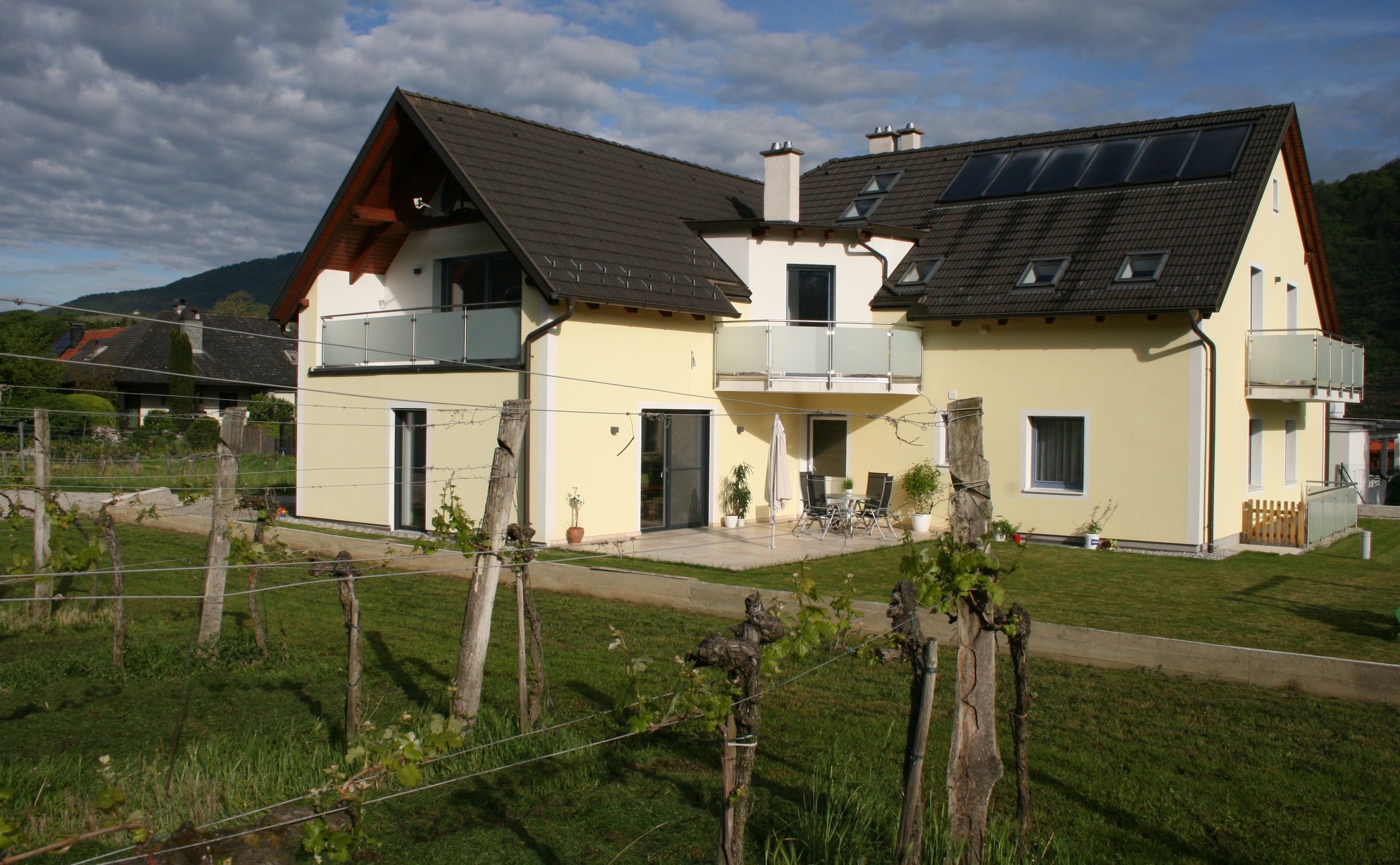 Yellow house with balcony and garden, surrounded by vines.