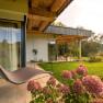 A modern vacation home with wooden veranda, hanging chair and blooming hydrangeas in the foreground, surrounded by green nature.